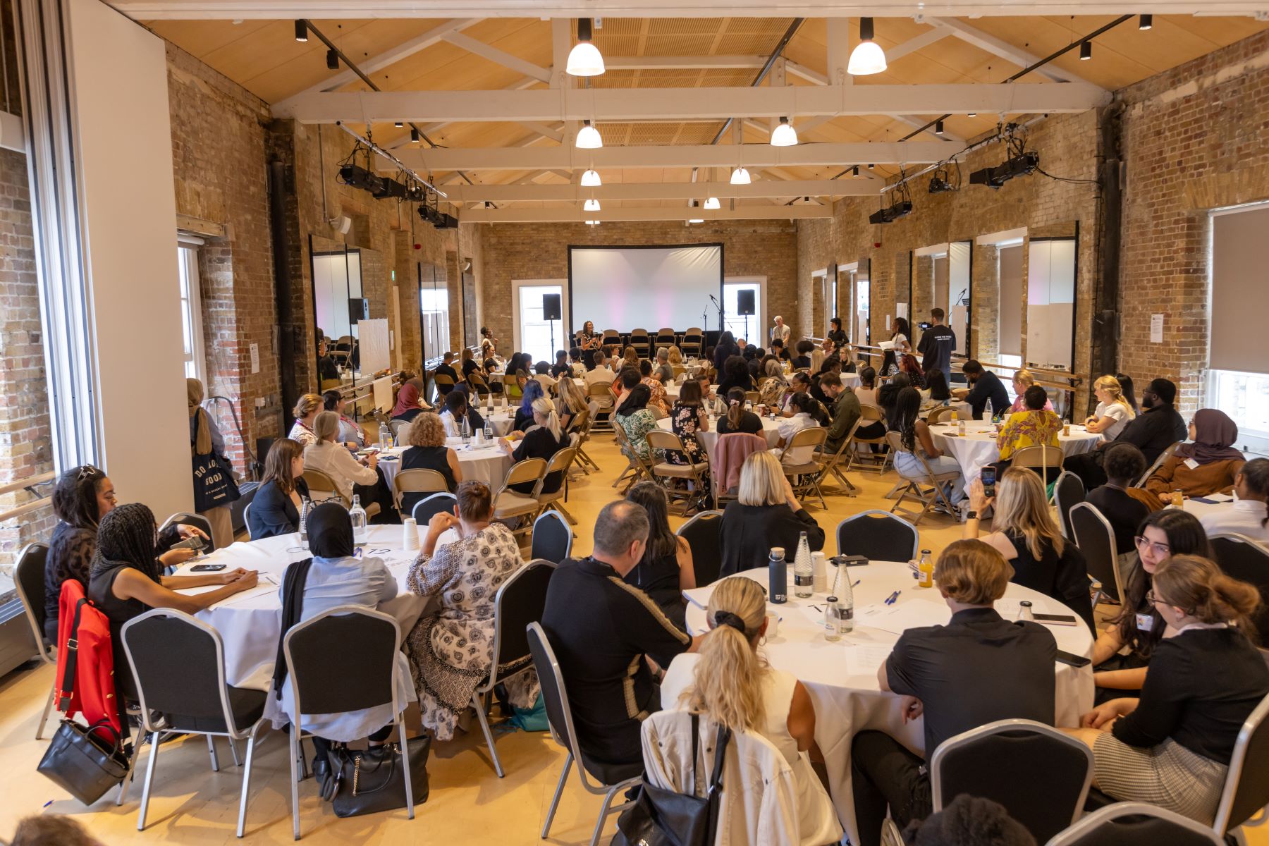 Photo from the back of the room at the Summit showing room filled with tables with 6-8 people sat at each one.